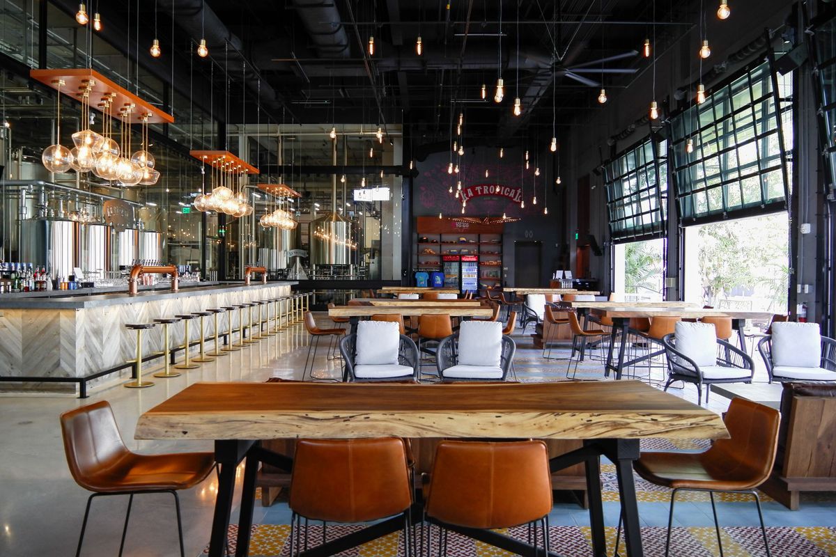 Cervecería con sillas color canela, mesa de madera, sillas blancas en el fondo y barra de hormigón